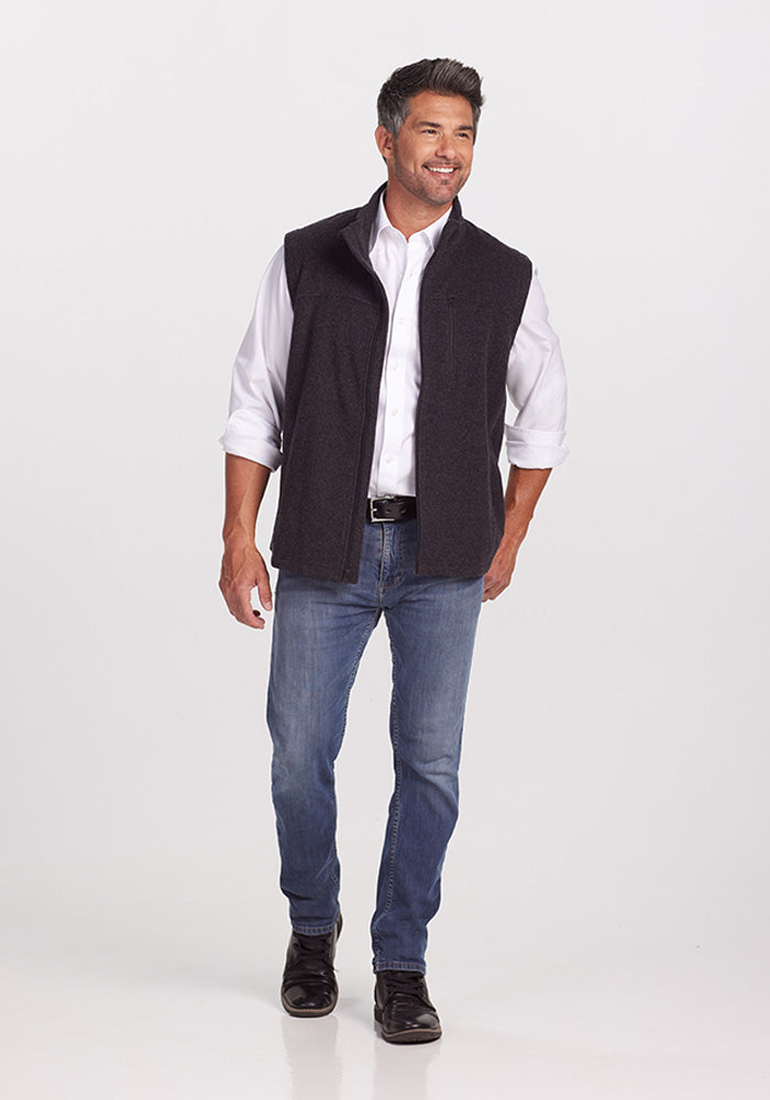 A man with short dark hair is standing and smiling against a plain white background. He is dressed in a white collared shirt with the sleeves rolled up, a Stowe Vest in Carbon Black from Woolx crafted from ultra-heavyweight Merino wool, blue jeans, and black boots.