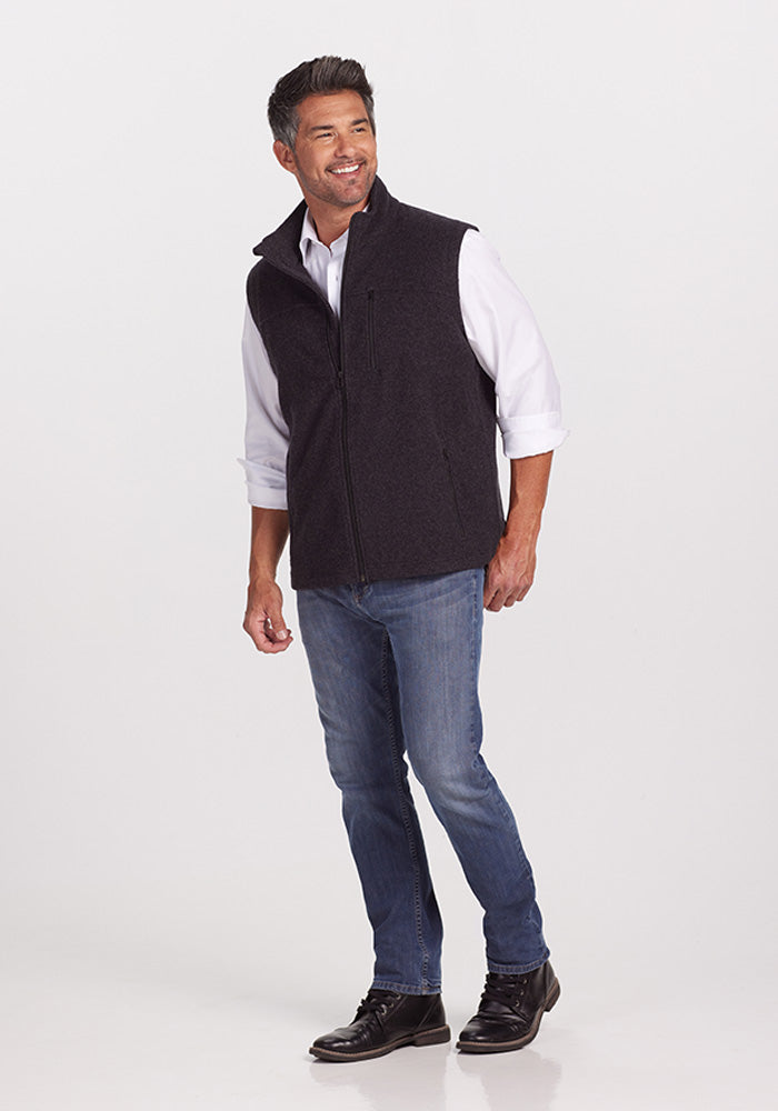 A man stands smiling against a plain white background. He is wearing a white shirt, the Carbon Black Stowe Vest from Woolx, blue jeans, and black lace-up shoes. The man has short dark hair and facial stubble. His body is slightly turned to the side, and one hand rests near his hip.