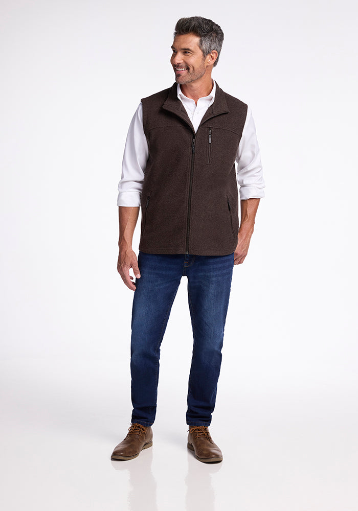 A man stands on a white background, wearing a white button-up shirt under a brown Stowe Vest from Woolx, blue jeans, and brown shoes. He has short dark hair, is smiling, and has one hand in his pocket.