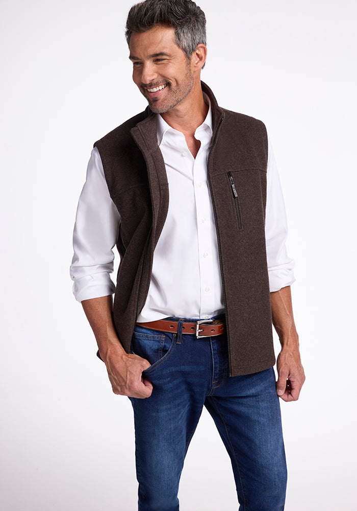 A man with salt-and-pepper hair smiles while wearing a white dress shirt, blue jeans, and the dark brown Stowe Vest by Woolx. He has one hand in his pocket and is posed against a plain white background.