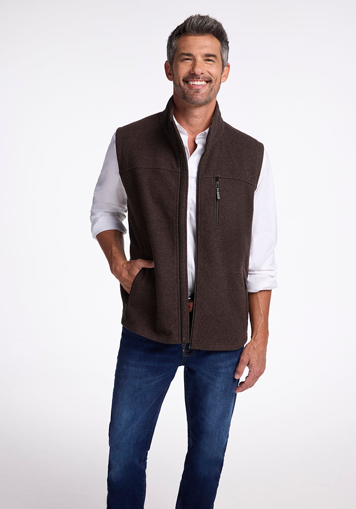 A person with short dark hair and a light beard smiles while standing against a plain background. They wear a white long-sleeve shirt, blue jeans, and the ultra-heavyweight Stowe Vest by Woolx that's perfect as a cold weather layer. Their hands are in their vest pockets. 