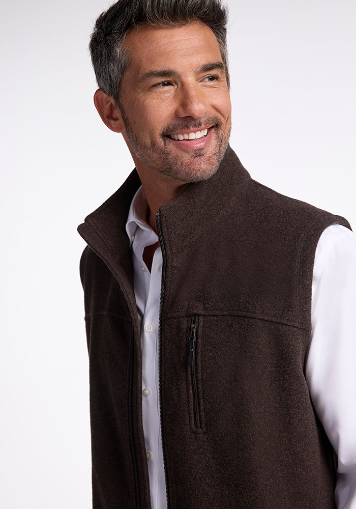 A man with short, dark hair and some gray streaks is smiling and looking to his right. He is wearing a white shirt and the Stowe Vest by Woolx, made of brown Merino wool featuring a zippered chest pocket, making it an ideal layer for cold weather. The background is plain white.