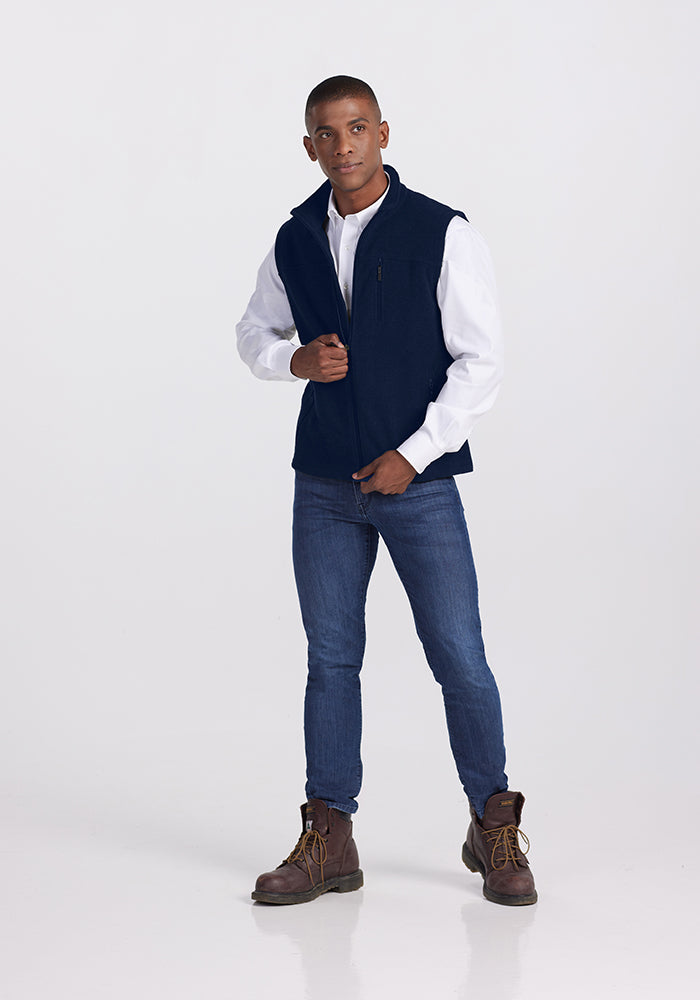 A person stands against a plain background, wearing a white long-sleeve shirt, the Woolx Stowe Vest in Deep Navy, blue jeans, and brown work boots. They have short hair and are adjusting the vest zipper with their left hand. Their gaze is directed towards the camera.