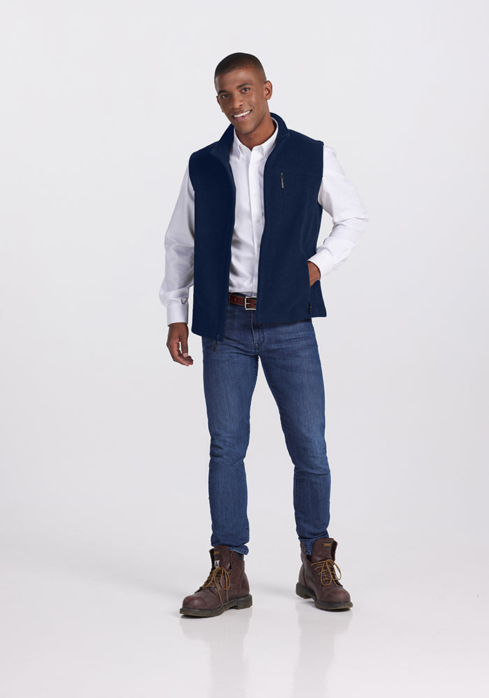 A person stands against a plain white background, wearing a white dress shirt, the Woolx Stowe Vest in Deep Navy made from ultra-heavyweight Merino wool, blue jeans, and brown lace-up boots. They have a relaxed posture, with one hand in their pants pocket and a slight smile on their face.