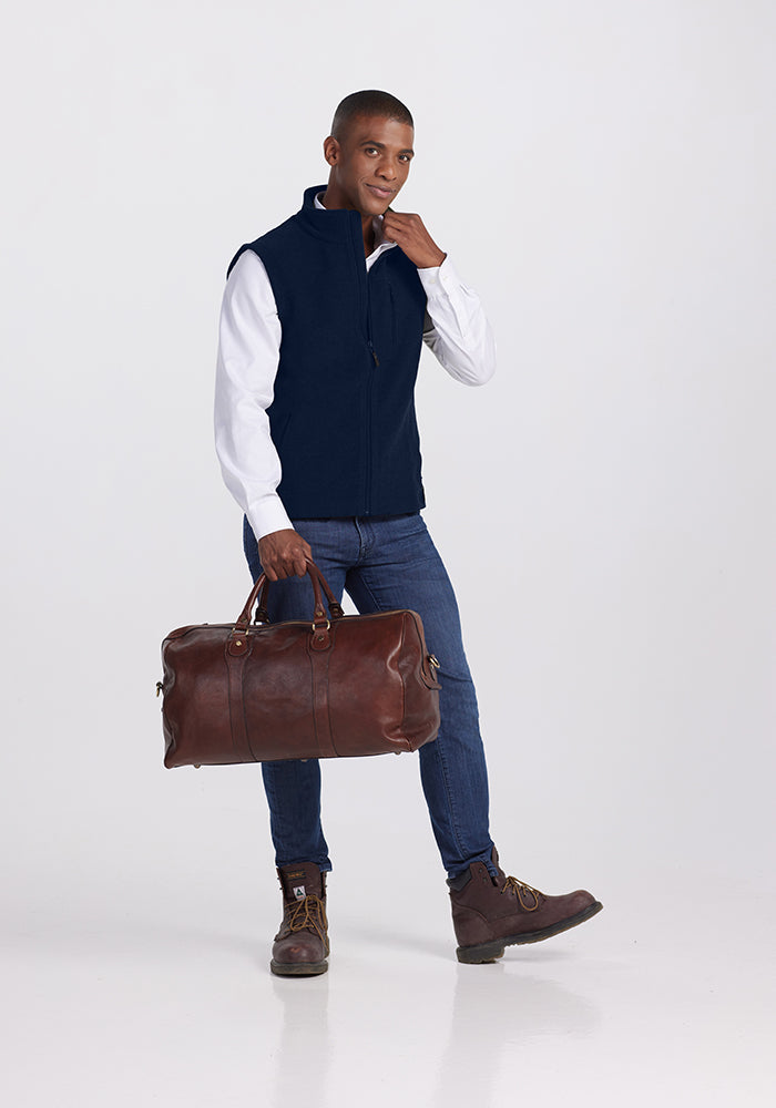 A person wearing a Woolx Stowe Vest in Deep Navy over a white shirt, blue jeans, and brown boots poses holding a brown leather duffel bag by its handles. The background is plain and light-colored.