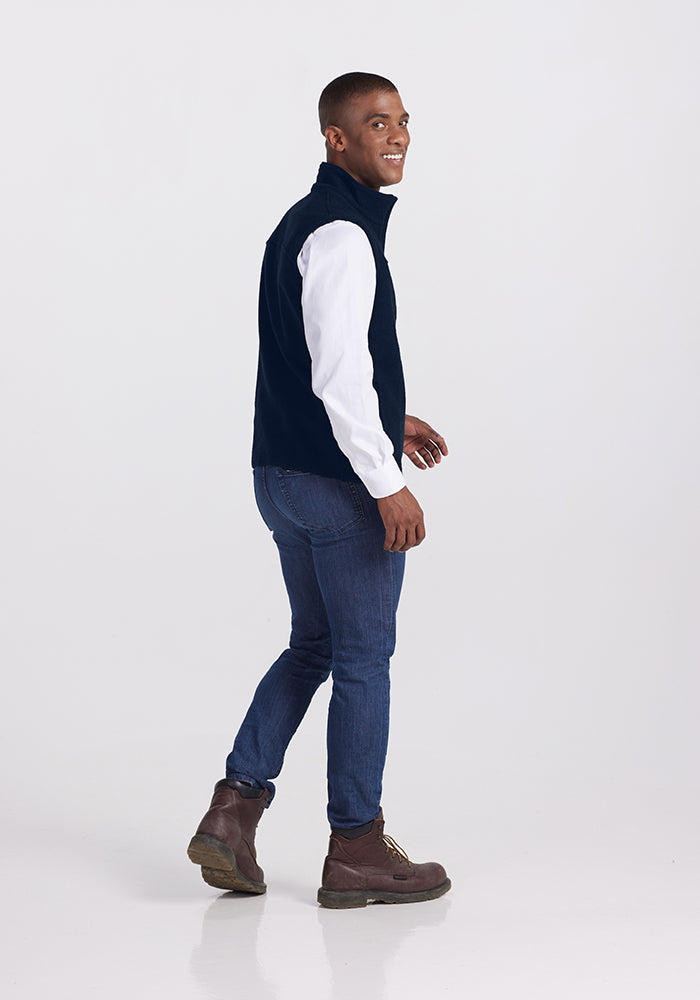 A man stands against a plain white background, looking off to the side with a smile. He is wearing an ultra-heavyweight, Merino wool Stowe Vest in Deep Navy by Woolx over a white long-sleeve shirt, complemented by blue jeans and brown boots. Captured in a slightly turned mid-walking pose, he epitomizes versatile outdoor gear.