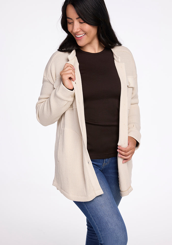 A person with long dark hair smiles while wearing the Woolx Oaklynn Waffle Shacket in oatmeal over a dark shirt and blue jeans. They are standing against a plain white background, holding the moisture-wicking shacket open with one hand.