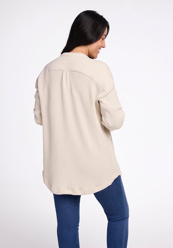 A woman with long dark hair, wearing the Oaklynn Waffle Shacket in oatmeal by Woolx and blue jeans, is turned slightly to the side, smiling against a plain white background.