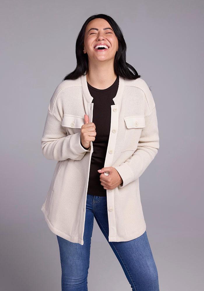 A person with long dark hair is wearing the Oaklynn Waffle Shacket in oatmeal by Woolx over a black shirt and blue jeans. They are smiling and laughing with their eyes closed, standing against a plain gray background.