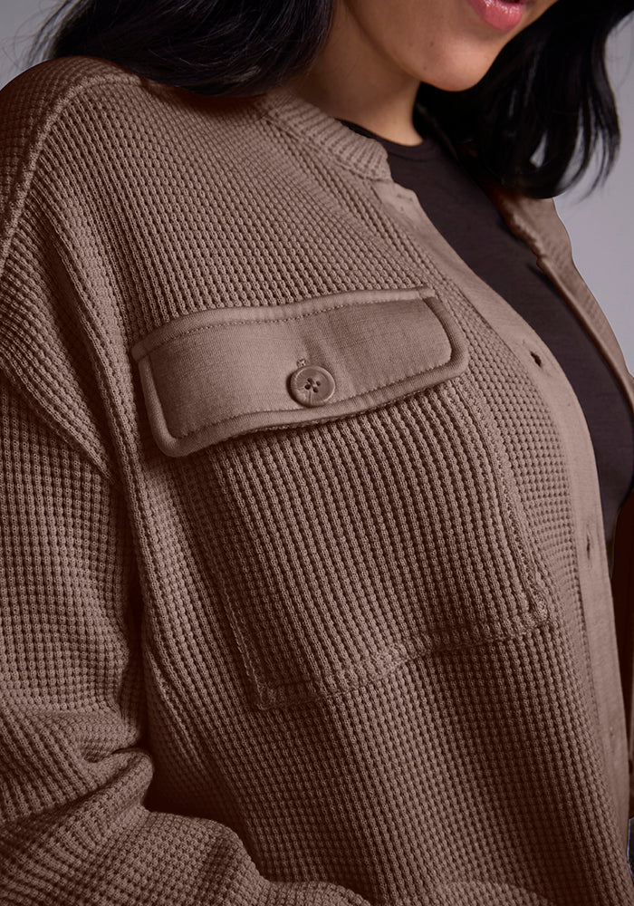 Close-up of a person wearing a textured brown Oaklynn Waffle Shacket - Latte by Woolx with a buttoned chest pocket over a black top. The image focuses on the intricate details and texture of the shacket, highlighting its unique design.