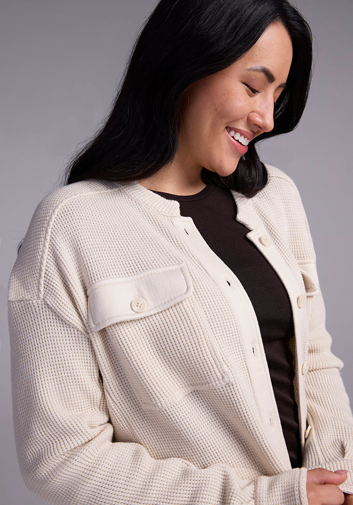 A woman with long black hair smiles while looking down, wearing the Woolx Oaklynn Waffle Shacket in oatmeal, crafted from soft merino wool, over a dark shirt. The neutral gray background enhances the cozy yet stylish vibe of her outfit.