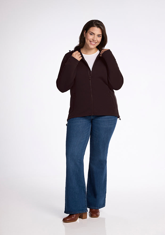 A person with shoulder-length dark hair smiles while wearing a black Woolx Cubby Hooded Sweatshirt over a white shirt, blue jeans, and brown ankle boots. They are standing against a white background and holding the hoodie collar adorned with heavy-duty zippers.