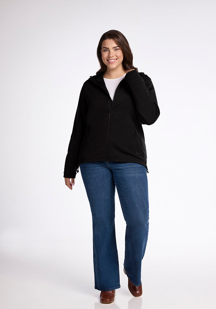 A woman stands against a plain white background, smiling. She is wearing a Woolx Cubby Hooded Sweatshirt - Black over a white shirt, blue jeans, and brown shoes. One hand rests by her side while the other touches her hair.