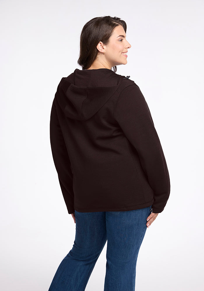 A person with shoulder-length hair is shown from the back, wearing a French Roast Woolx Cubby Hooded Sweatshirt and blue jeans. They are looking to the side with a slight smile and have one hand resting on their hip. The background is plain white.