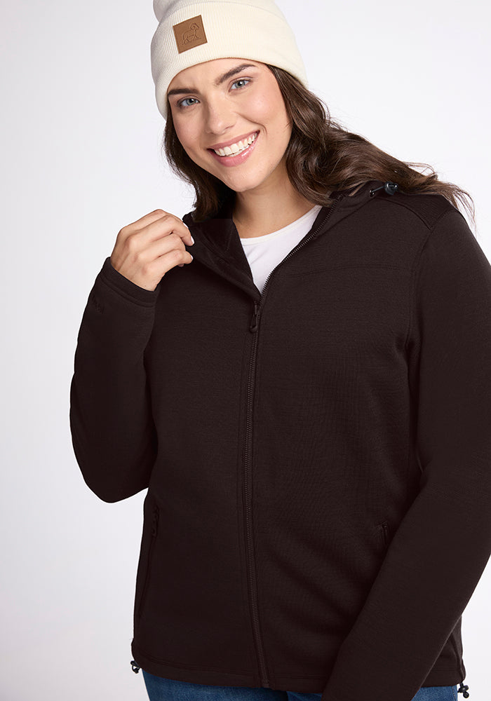 A woman with long dark hair smiles while wearing a French Roast Cubby Hooded Sweatshirt by Woolx and a white beanie. The background is plain white, and the woman appears relaxed and happy.