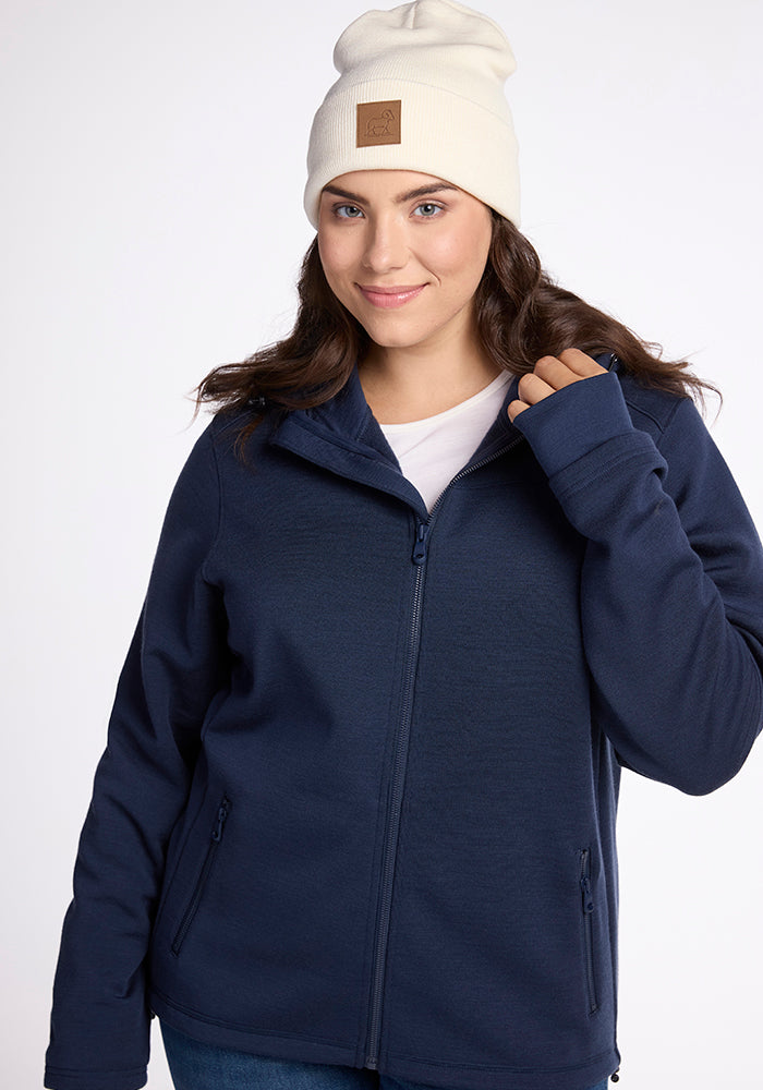 A person with long hair is wearing a Woolx Baylor Beanie in white, a navy blue zip-up jacket, and a white shirt. They are smiling and holding onto their jacket's zipper with one hand. The background is plain white.