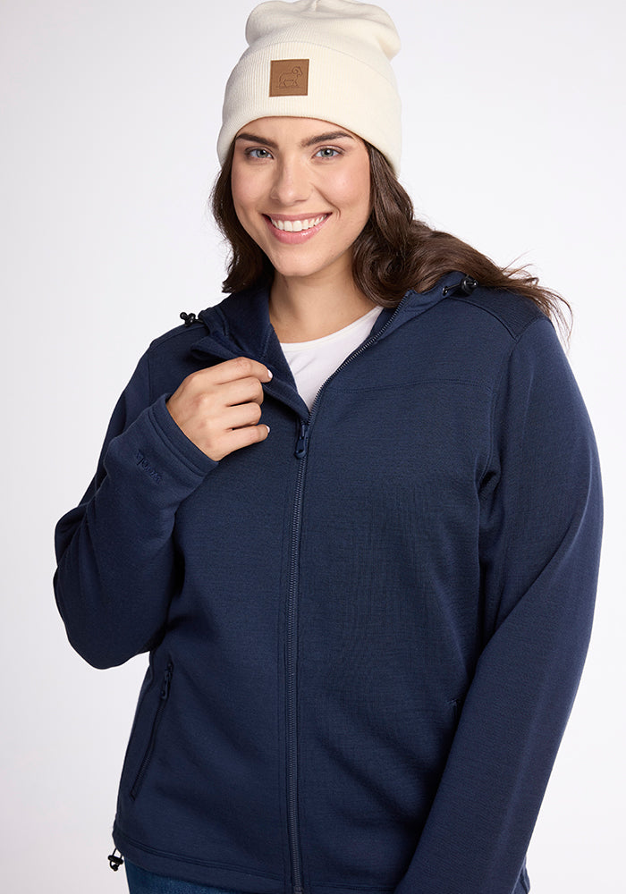 A person is smiling and looking at the camera while wearing a white Baylor Beanie by Woolx and a navy blue zip-up hoodie. The background is plain and light-colored.