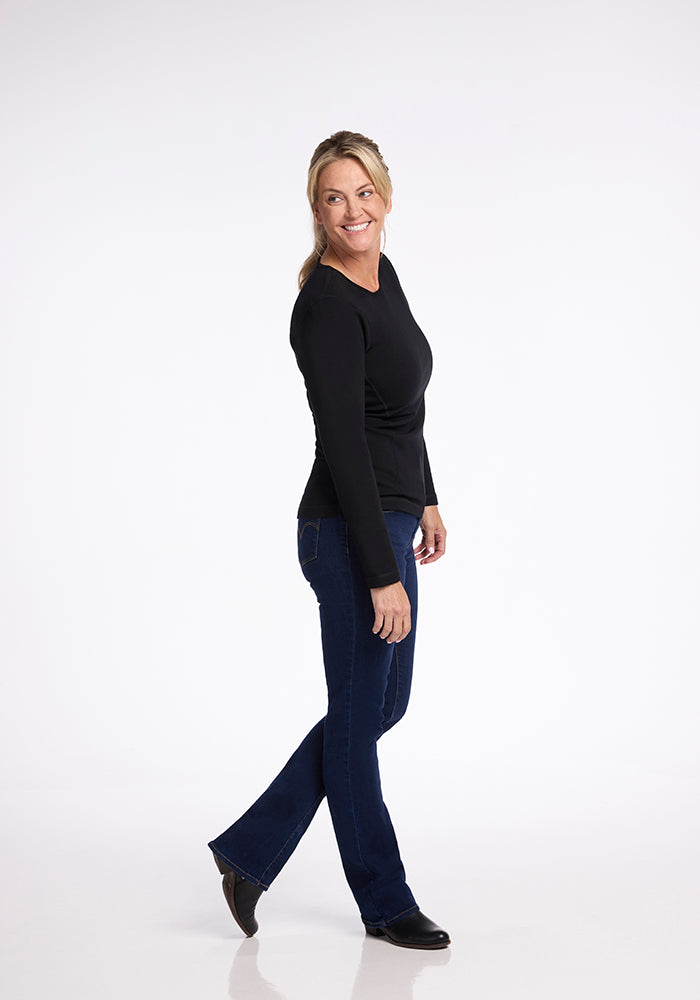 A person with long blond hair is smiling and standing sideways, wearing the heavyweight Riley Long Sleeve Crew OLD by Woolx, paired with dark blue jeans and black shoes. The background is plain white.