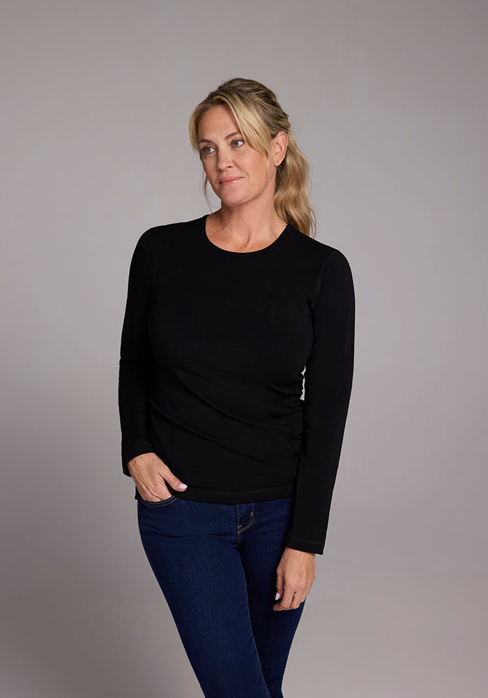 A person with long, blonde hair is wearing the Riley Long Sleeve Crew in black from Woolx and blue jeans. They stand against a plain gray background, looking slightly to the side with a calm expression.