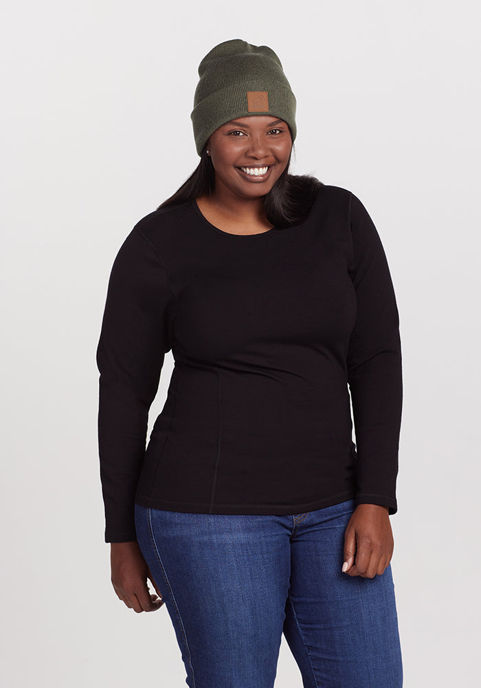 A person is smiling while standing against a plain background. They are wearing a black, long-sleeved shirt, blue jeans, and a green Baylor Beanie - Spruce Heather from Woolx with a patch on the front.