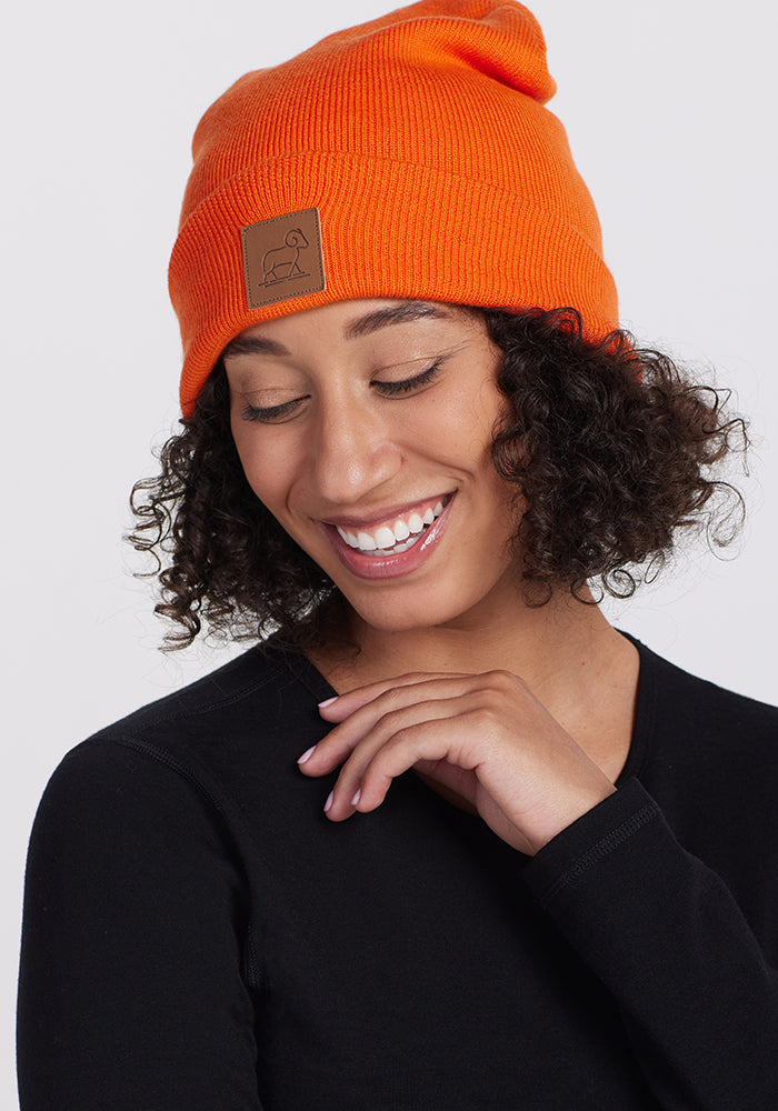 A person with curly hair is smiling while looking down. They are wearing a bright orange Baylor Beanie made from Australian merino wool by Woolx, which features a brown patch, and they have on a black long-sleeve shirt.