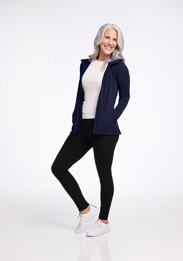 An older woman with long gray hair smiles while standing against a plain white background. She is wearing a white top, the Woolx Zoey Zip Up Hoodie in Deep Navy made of DuraLite Fabric, black pants, and white sneakers. Her hands rest in the pockets of her hoodie as she leans slightly to one side.