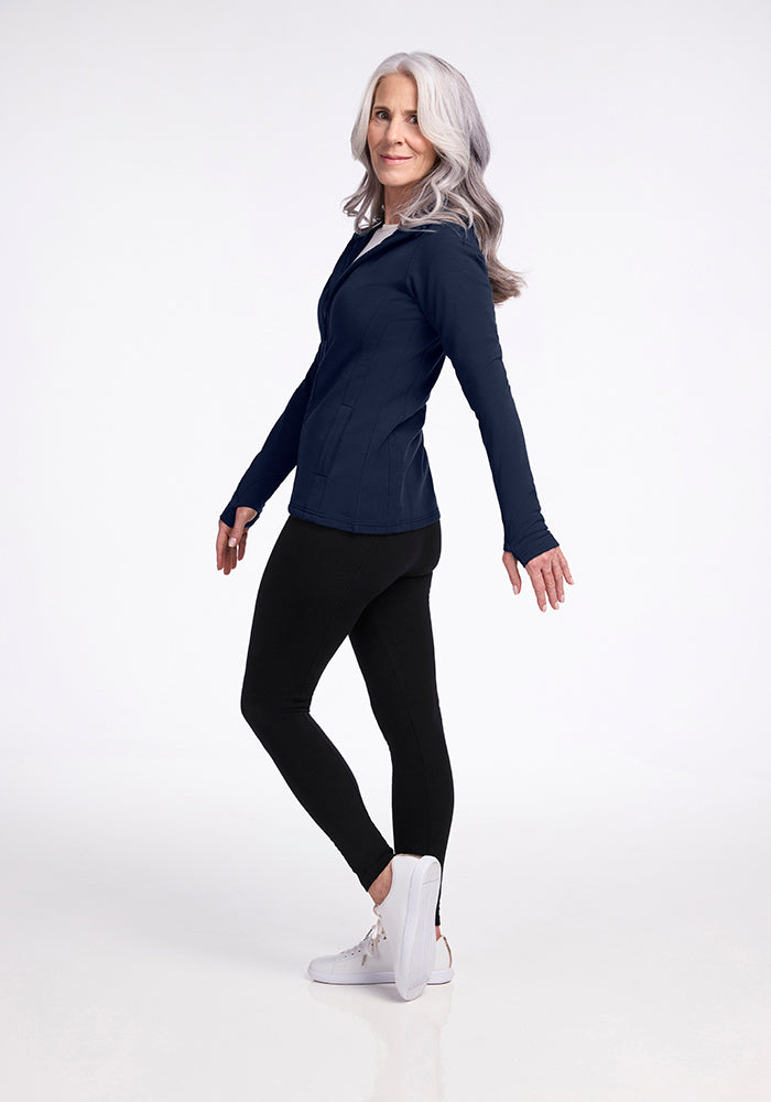 A woman with long, wavy gray hair is posing in a deep navy Woolx Zoey Zip Up Hoodie crafted from DuraLite Fabric, paired with black leggings and white sneakers. She is slightly turned to the side with her arms bent at the elbows and a slight smile on her face. The background is plain white.