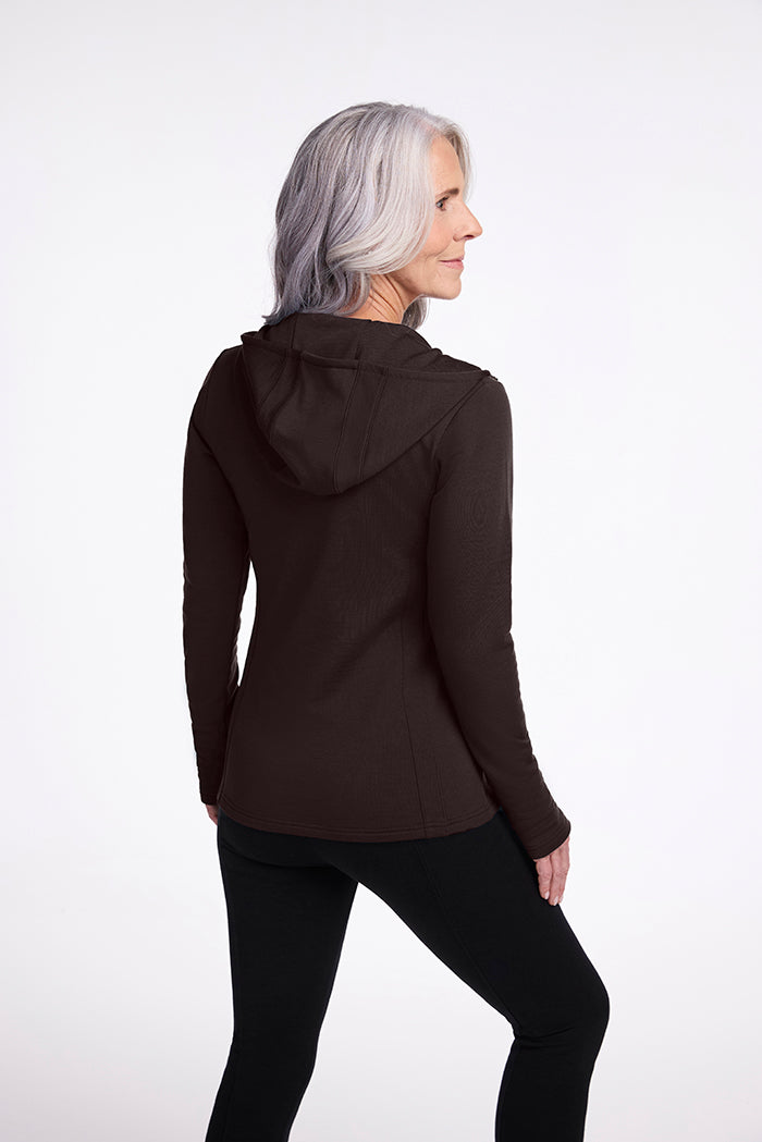 A woman with long gray hair stands facing away, turning slightly to the side. She is wearing the Zoey Zip Up Hoodie in French Roast from Woolx, made from soft Australian Merino Wool, along with black pants. The background is a plain white.