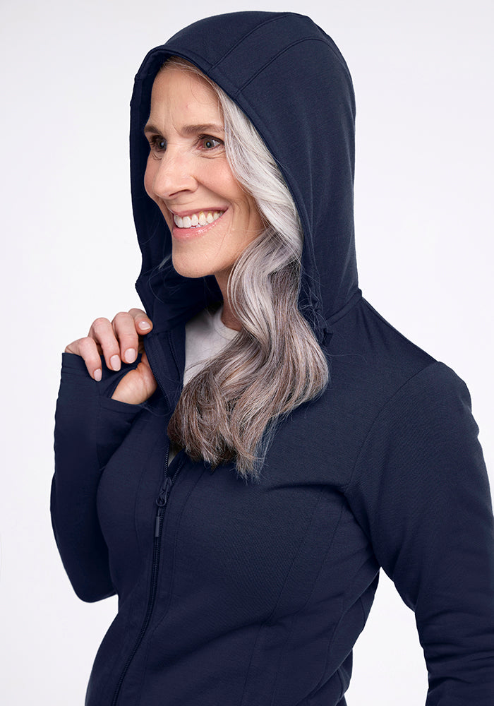 A woman with long, wavy gray hair smiles while holding the edge of the hood on her deep navy Woolx Zoey Zip Up Hoodie. Made from Woolx DuraLite Fabric, she faces to the left, wearing a white shirt underneath. The background is plain and light-colored.