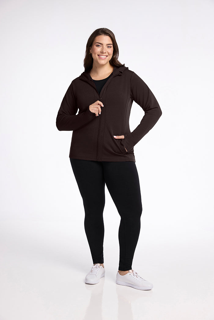 A person with long, dark hair is standing and smiling at the camera. They are wearing a black Zoey Zip Up Hoodie - French Roast from Woolx made from Australian Merino Wool, black leggings, and white sneakers. The background is plain and white.