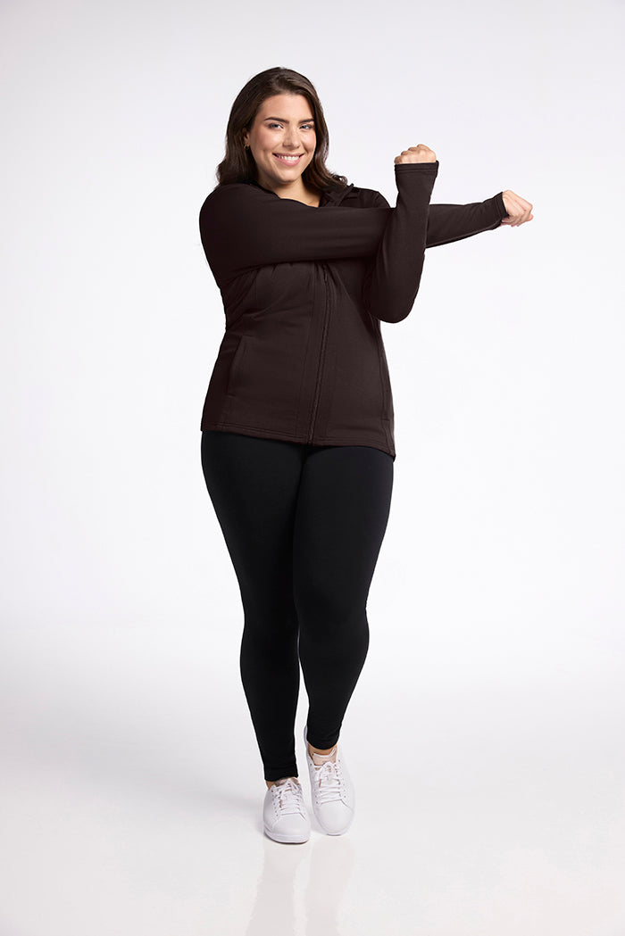 Stylishly slim Zoey, wearing the Woolx Zoey Zip Up Hoodie in French Roast, black leggings, and white sneakers, stands against a plain white background. She stretches her arm across her chest and smiles brightly.