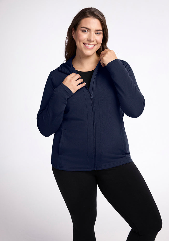 A woman with long dark hair smiles and stands confidently, one hand holding the collar of her deep navy Zoey Zip Up Hoodie from Woolx, made from Australian Merino Wool. She is dressed in black leggings and a black shirt, posing against a plain white background. 