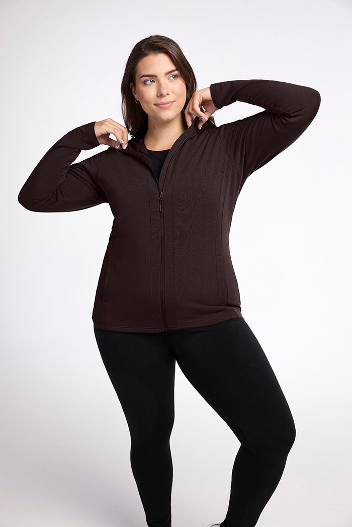 A person with long, brown hair is modeling a dark brown Woolx Zoey Zip Up Hoodie - French Roast and black leggings. They have their hands raised, holding the hood with a subtle smile on their face. The background is plain white. 