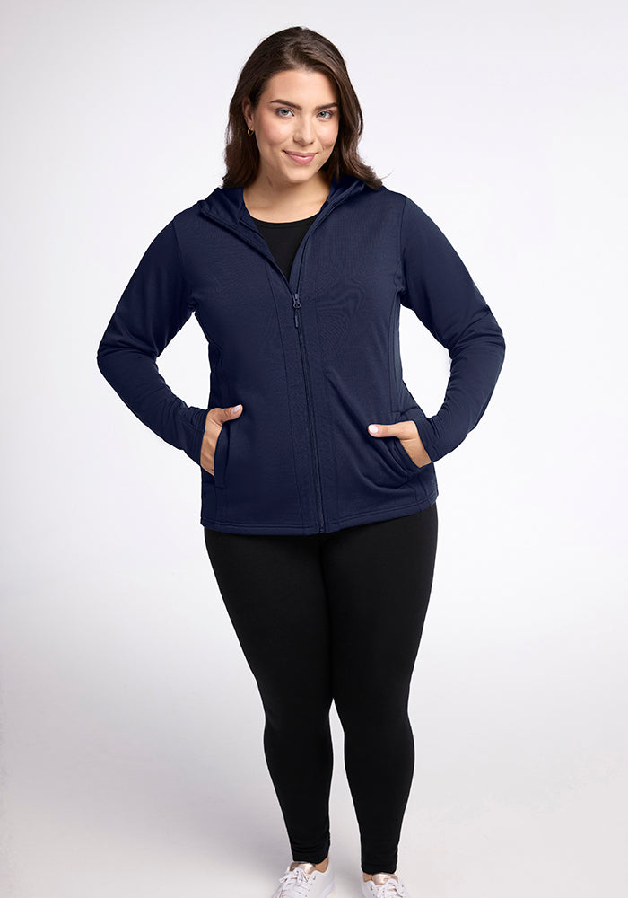 A woman is standing against a plain background, wearing a deep navy Zoey Zip Up Hoodie from Woolx made from Australian Merino Wool, paired with black leggings and white sneakers. Her hands are in the pockets of the mid-weight hoodie, and she is smiling at the camera.