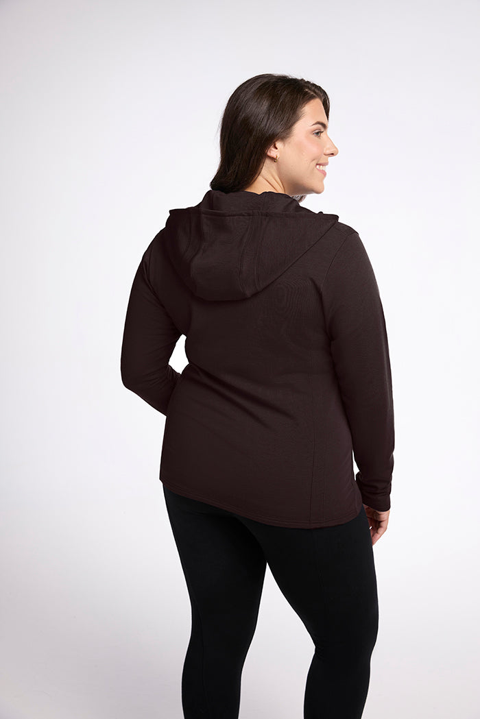 A woman is shown from the back, wearing a dark-colored Zoey Zip Up Hoodie - French Roast by Woolx and black leggings. She has long, dark hair and is smiling slightly. The background is plain and white. The stylishly slim Zoey design highlights her silhouette gracefully.