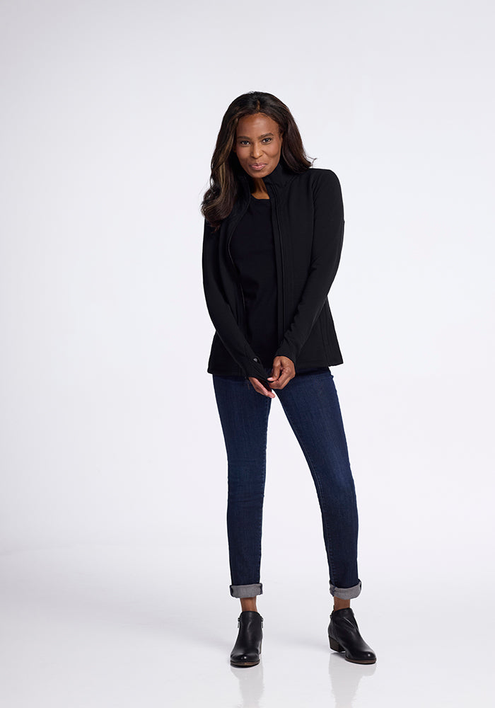 A woman with long hair, dressed in a Woolx Finley Zip Up Sweatshirt - Black over an Australian merino wool shirt and dark blue rolled-cuff jeans, stands against a plain white background. She is wearing black ankle boots and has a gentle smile, creating a flattering look.