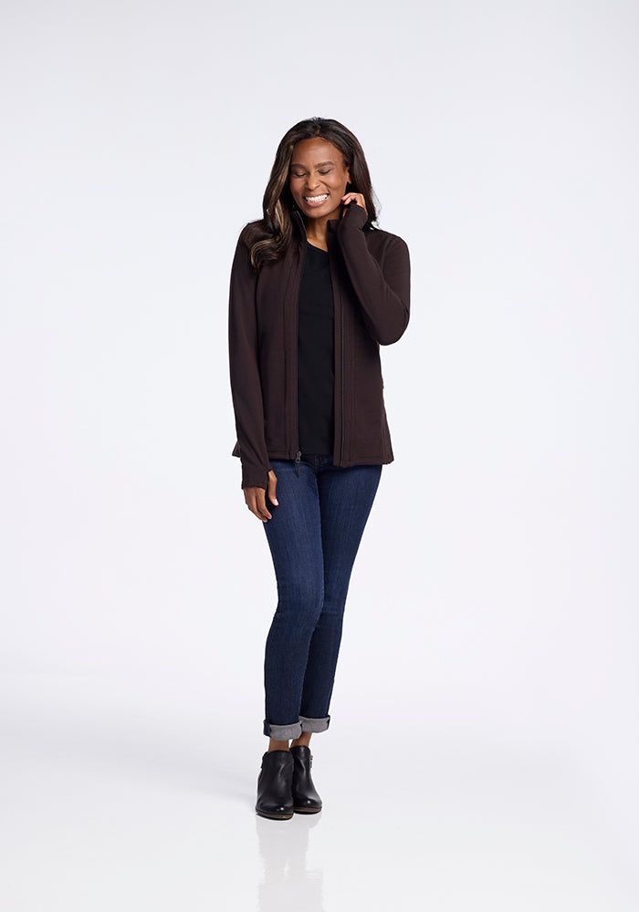 A woman stands against a plain background, smiling with one hand touching her cheek. She is wearing the Finley Zip Up Sweatshirt in French Roast by Woolx made of Australian merino wool over a black top, dark blue jeans, and black ankle boots. She has long, straight hair and appears relaxed and confident.