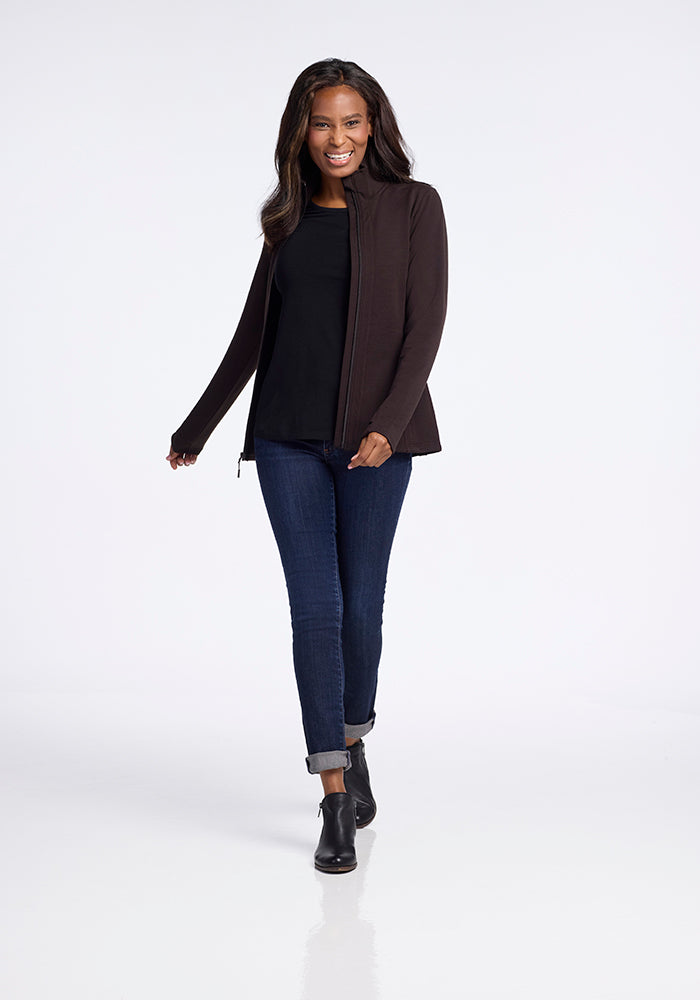 A woman with long dark hair walks confidently in a light-filled space. She is wearing a black jacket over a Finley Zip Up Sweatshirt in French Roast by Woolx, dark blue cuffed jeans, and black ankle boots. She is smiling and appears relaxed and happy.