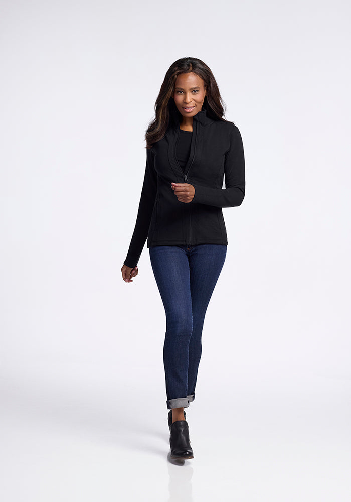 A woman with long hair is walking forward, wearing the Woolx Finley Zip Up Sweatshirt in black, dark blue jeans, and black ankle boots. The background is a plain light grey. She has a confident expression and posture.