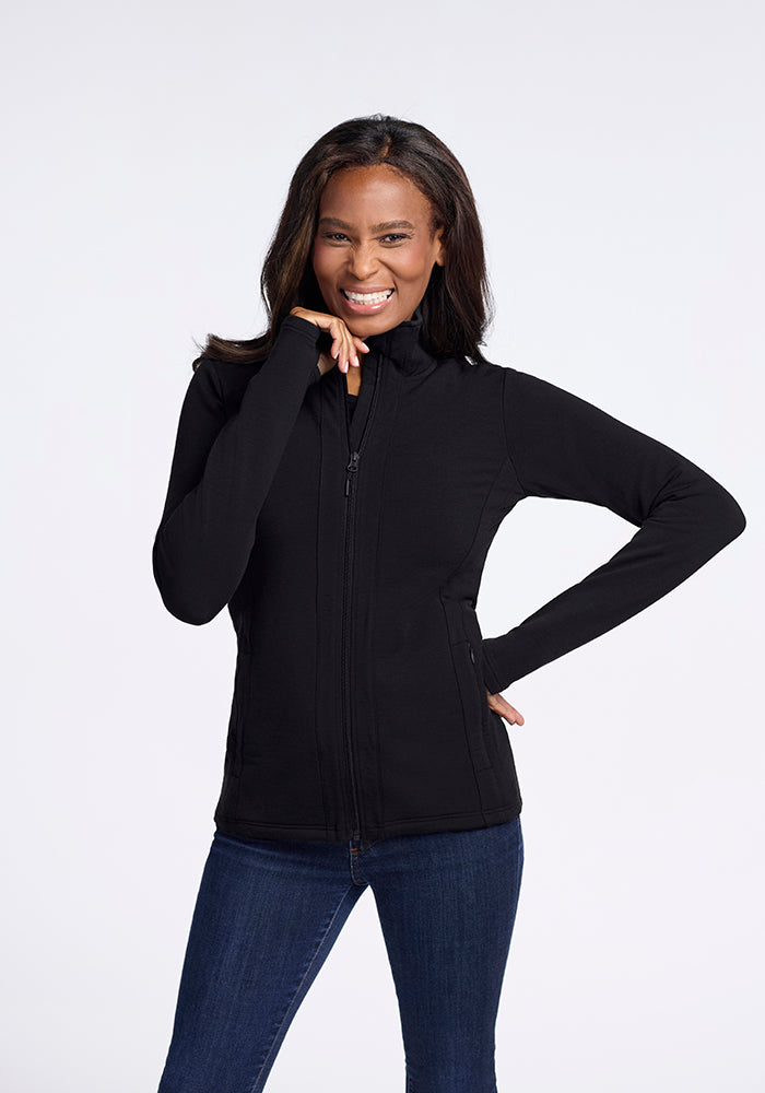 A woman with long hair is smiling and posing against a plain white background. She is wearing the Finley Zip Up Sweatshirt in black from Woolx, made of luxurious Australian merino wool, paired with blue jeans. With her left hand on her hip and her right hand touching her face, she effortlessly showcases the flattering look provided by the no-hood design of the sweatshirt.