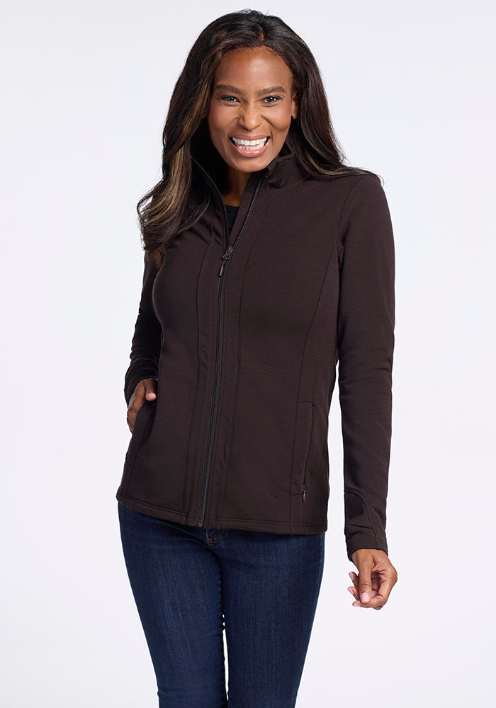 A woman with long, dark hair smiles widely while posing in the stylish Finley Zip Up Sweatshirt - French Roast by Woolx and dark jeans. She stands against a plain white background, with her left hand in her jacket pocket and her right hand relaxed by her side.