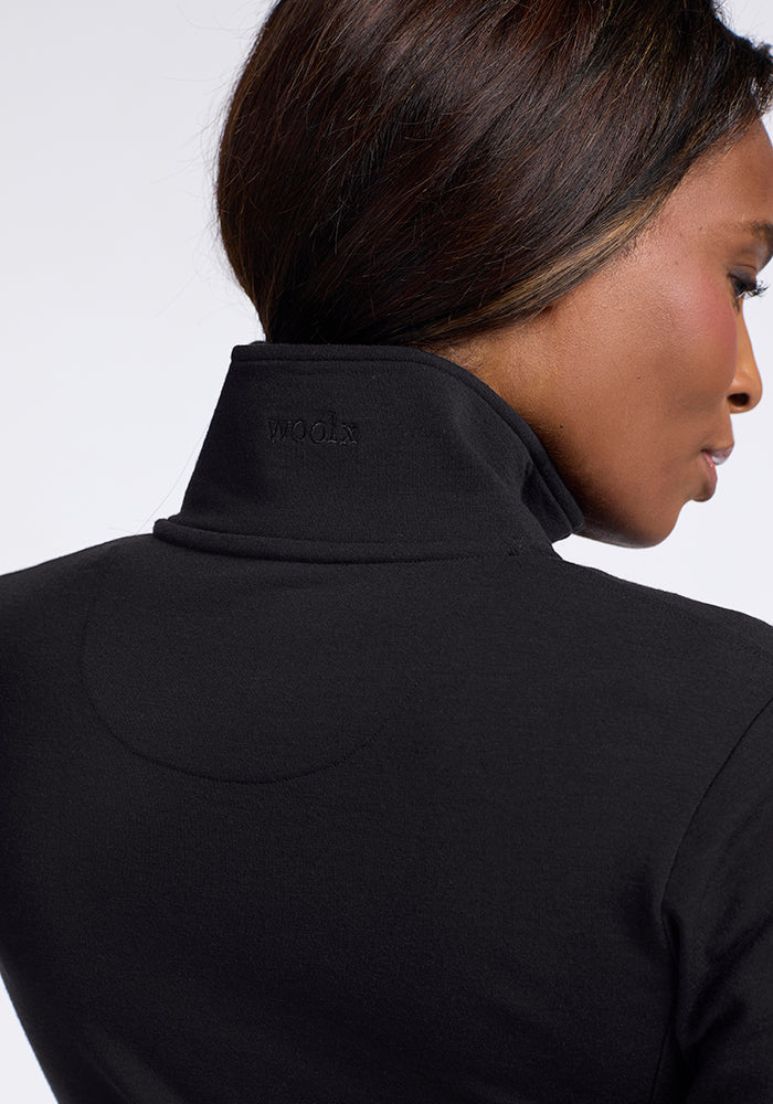 A person with dark hair is shown from the back and side, wearing a flattering black Finley Zip Up Sweatshirt from Woolx, subtly stitched with the word "KNOW" into the collar. Made from premium Australian merino wool, this stylish piece stands out prominently against the neutral background.