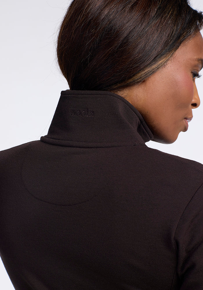 A close-up image of a person with long dark hair from behind, showcasing the upper back and neck. They are wearing a black Finley Zip Up Sweatshirt in French Roast by Woolx, made from Australian merino wool, with the word "xxom." subtly embroidered near the collar. The background is white.