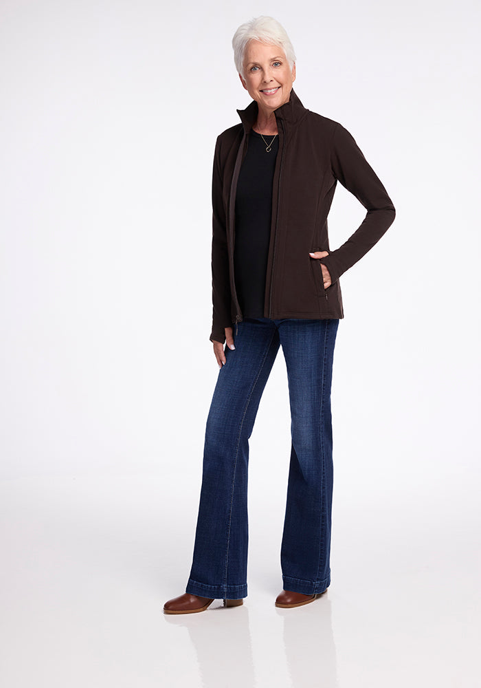 An older adult with short white hair stands and smiles with their hands in the pockets of a Finley Zip Up Sweatshirt in French Roast by Woolx. They wear a black shirt, blue jeans, and brown shoes made from Australian merino wool against a plain white background.