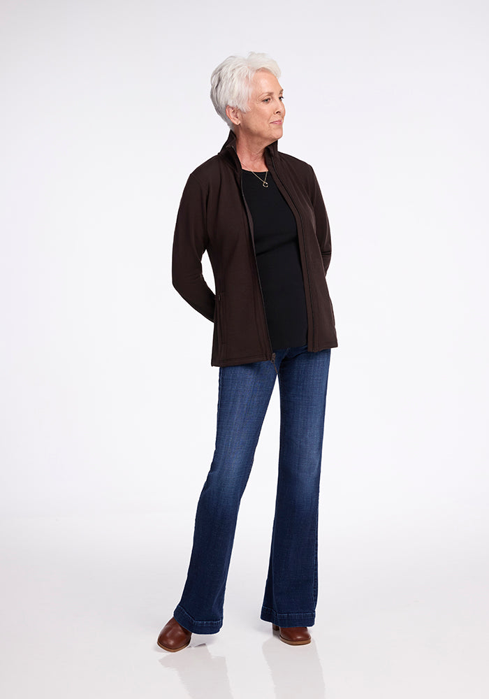 An older adult with short white hair stands against a plain white background. They wear a black top under a dark Australian merino wool Finley Zip Up Sweatshirt in French Roast by Woolx, blue jeans, and brown shoes. The person stands with their hands behind their back, gazing slightly to one side.