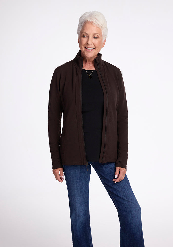 An older woman with short white hair is smiling and standing against a plain background. She is wearing a French Roast-colored Finley Zip Up Sweatshirt by Woolx over a black top and blue jeans made from Australian merino wool. Her hands are relaxed at her sides, exuding warmth like a fresh cup of French roast.