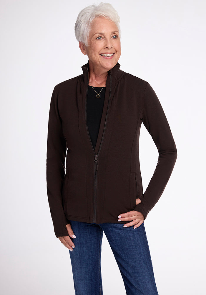 An older woman with short gray hair is smiling while wearing the Finley Zip Up Sweatshirt in French Roast by Woolx over a black top and blue jeans. The background is plain white.