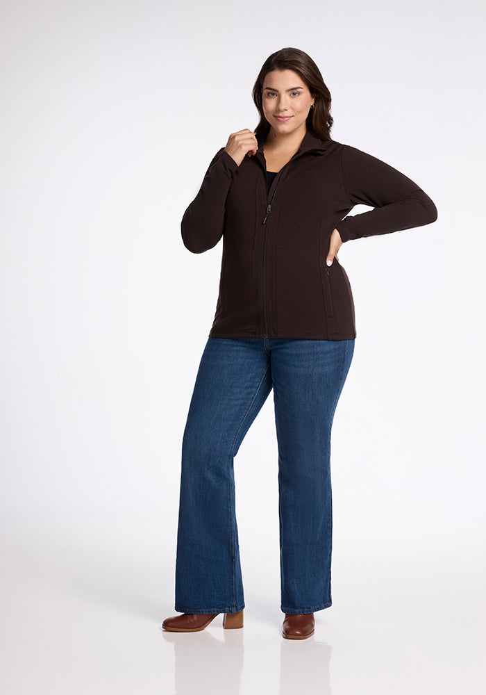 A woman stands confidently with a slight smile, posing in the Finley Zip Up Sweatshirt - French Roast by Woolx, paired with blue jeans and brown heeled shoes. She has one hand resting on her hip and the other partly raised towards her chin. The background is plain white.