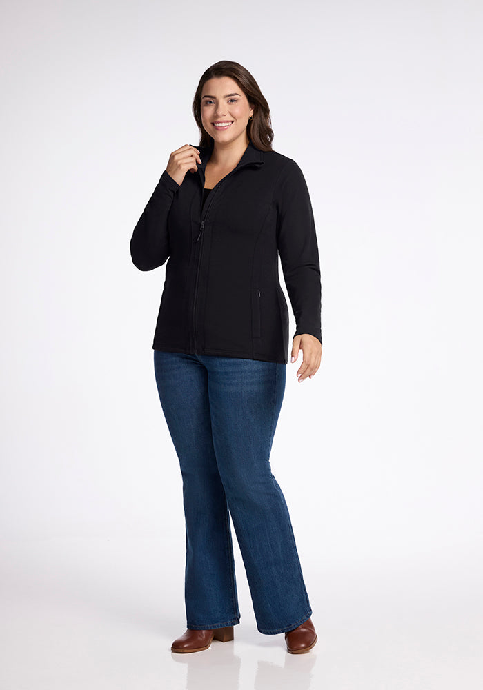 A person with shoulder-length dark hair smiles while facing forward. They are wearing a flattering, black Finley Zip Up Sweatshirt from Woolx, paired with blue jeans and brown shoes, standing in a well-lit space with a plain white background. One hand is raised to their collar, and the other hangs by their side.