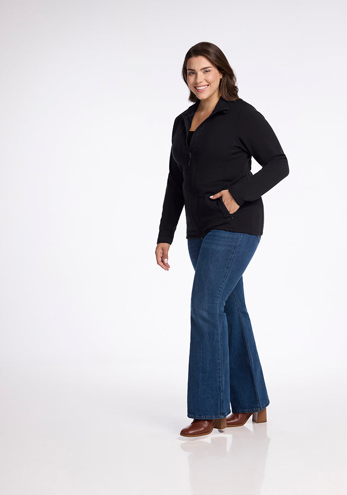 A woman with long dark hair is smiling and walking. She is wearing the Finley Zip Up Sweatshirt in black by Woolx, paired with blue flared jeans and brown heeled shoes. The flattering look is set against a plain white background.